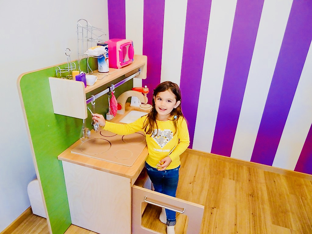 Une fillette se tient dans une pièce colorée devant une cuisine de jeu. Nous accordons une importance particulière à l'encouragement des capacités créatives des enfants, car celles-ci peuvent favoriser le développement de la personnalité. On peut exprimer sa créativité dans de nombreux domaines, que ce soit en peignant, en lisant ou, comme ici, dans la cuisine.