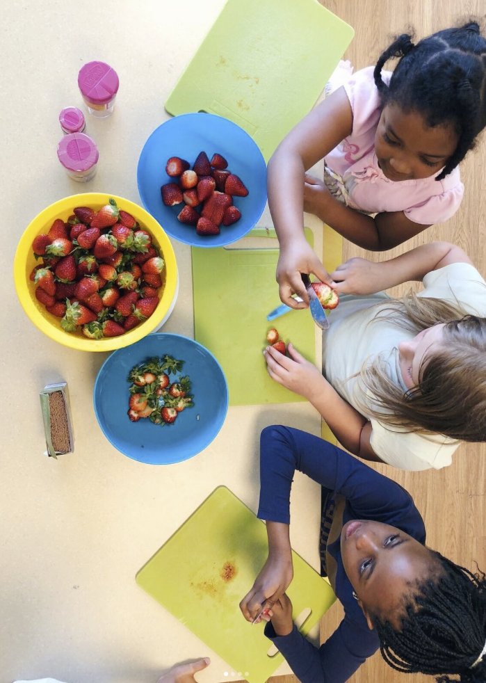 Trois enfants sont assis à une table. Devant eux se trouvent trois bols. L'un contient des fraises entières, l'autre des feuilles vertes et le troisième des fraises sans les feuilles vertes. Une alimentation saine est essentielle pour tout développement et joue un rôle primordial dans nos crèches.