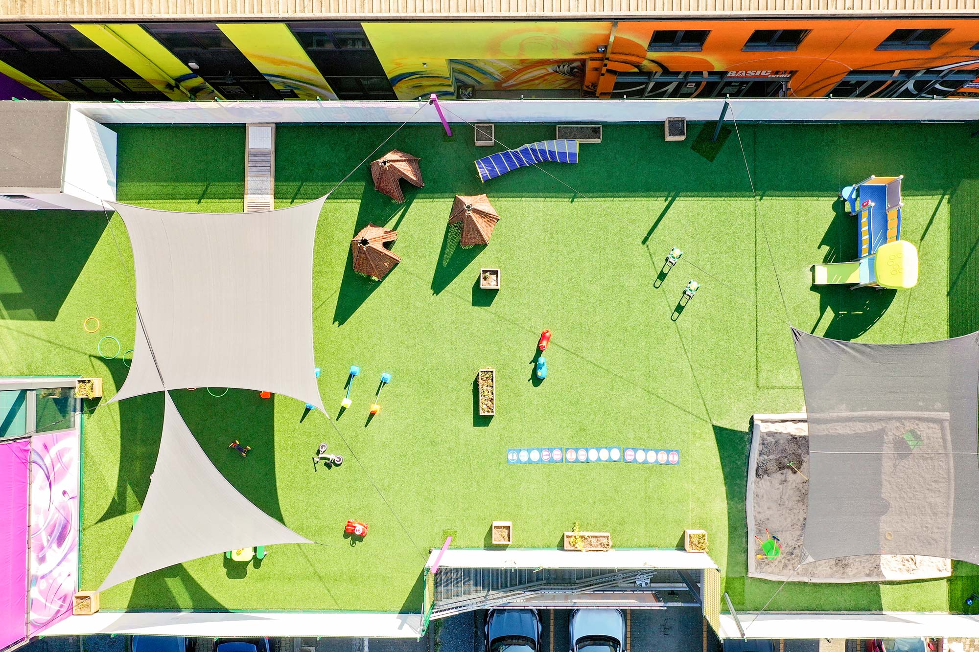 Vue à vol d'oiseau de la cour intérieure de notre crèche à Ettelbruck. Sur le sol vert, on voit divers modules de l'aire de jeux, dont des tentes Tippie, un château à escalader avec glissière et un bac à sable. Les différents éléments de nos espaces extérieurs ont comme but d'éveiller l'intérêt de chaque enfant afin qu'il puisse jouer dehors en toute sécurité.