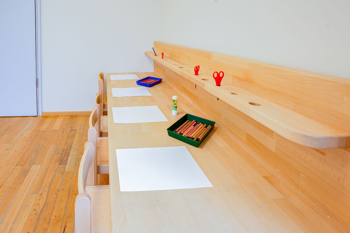 On y voit un banc en bois surmonté d'une table. Dans la table, il y a un rebord avec des trous ronds dans lesquels sont insérés des ciseaux. Nous choisissons nos équipements pour qu'ils fonctionnent selon la pédagogie Pikler, qui met l'accent sur le développement de l'autonomie des enfants.