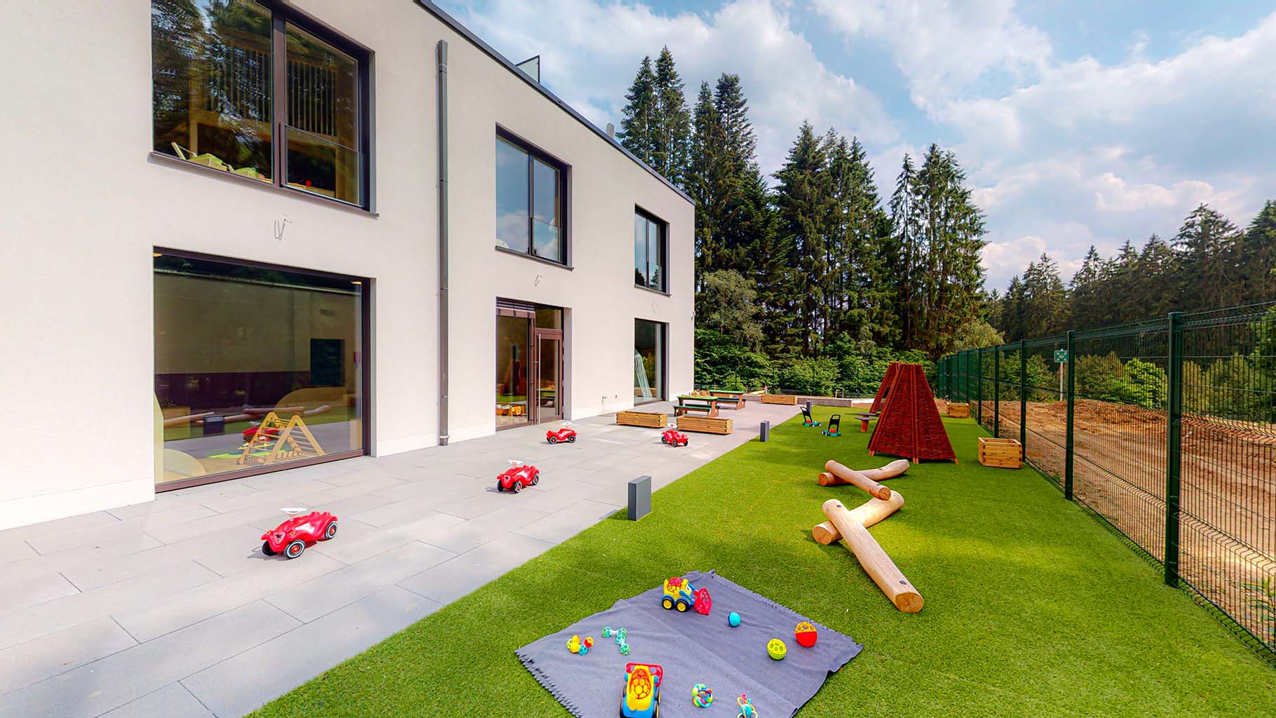 Une vue latérale de la crèche de Wemperhardt. On y voit un bâtiment moderne avec une grande surface de jeu. Sur celle-ci se trouvent quelques bobbycars, quelques poutres en bois qui se superposent pour entraîner l'équilibre. A l'avant de l'image, une couverture est étalée sur laquelle on peut voir deux petites pelleteuses et d'autres jouets. Plus loin, on voit deux tipies en bois. Ces espaces extérieurs favorisent le mouvement des enfants, de sorte qu'ils sont pleinement occupés.