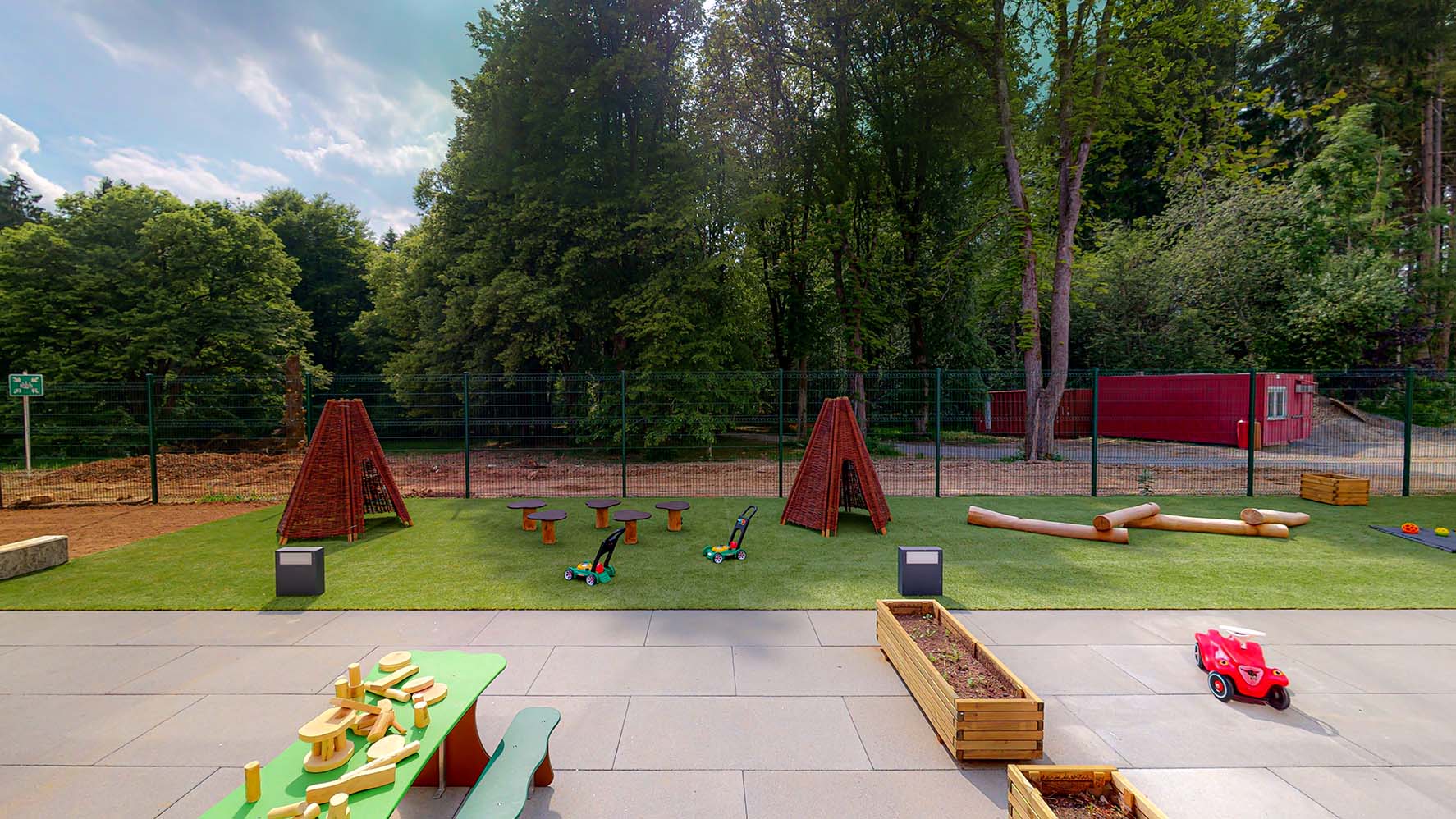 Sur la vue de face de l'espace extérieur de notre crèche à Wemperhardt, on voit une table de pique-nique sur laquelle on peut voir des jouets en bois, un parcours pour travailler l'équilibre et quelques tipies qui sont également en bois. Les divers équipements de nos espaces extérieurs motivent les enfants à passer beaucoup de temps à l'air libre.