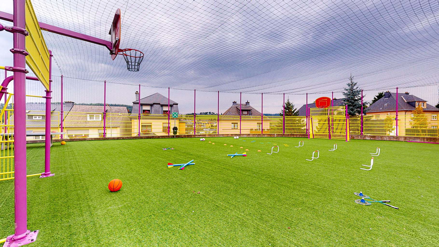 Le terrain de sport de Wiltz a un sol vert. Deux paniers de basket-ball et des buts de football sont visibles à gauche et à droite. On y voit également quelques fusées de jeu, des haies et d'autres éléments qui indiquent que des activités variées ont lieu. Un filet en tissu entoure le terrain de sport. Cet équipement garantit un quotidien varié aux enfants en toute sécurité.