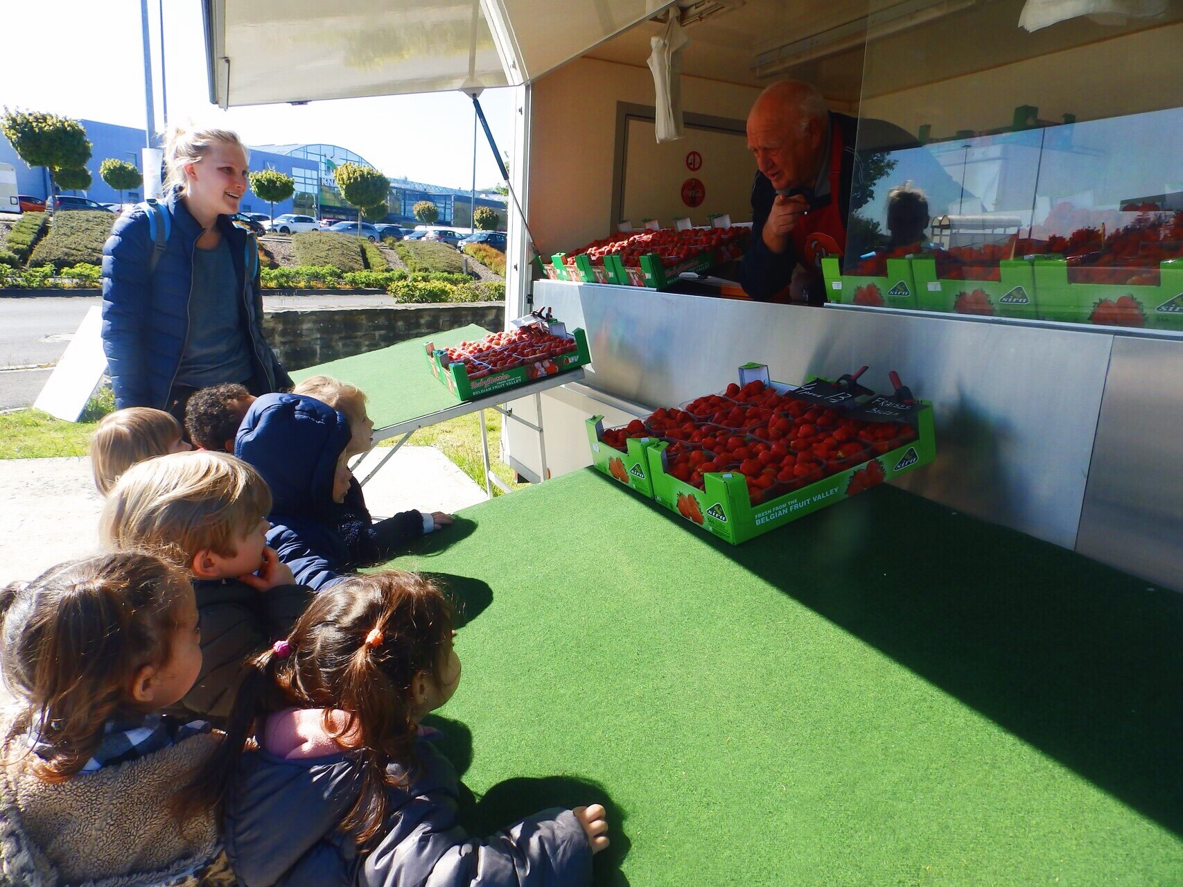 Une animatrice se tient à côté d'un groupe d'enfants devant un stand de marché qui ne vend que des fraises. Le vendeur regarde les enfants comme s'il leur expliquait quelque chose. Pour nous, il est important que les enfants sachent dès leur plus jeune âge ce que signifie une alimentation saine.