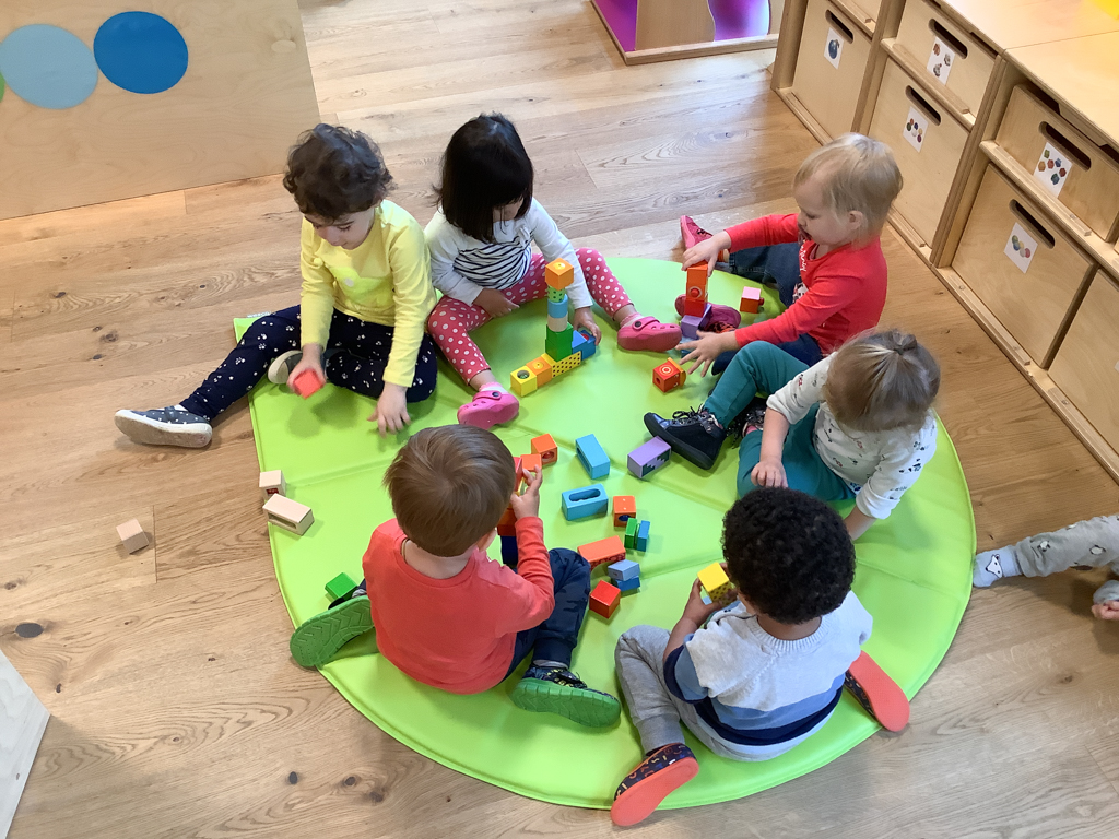 On y voit six enfants assis sur une surface verte et jouant ensemble avec des blocs de construction. Jouer ensemble favorise les compétences sociales et permet aux enfants de nouer de nouveaux contacts dans nos crèches.