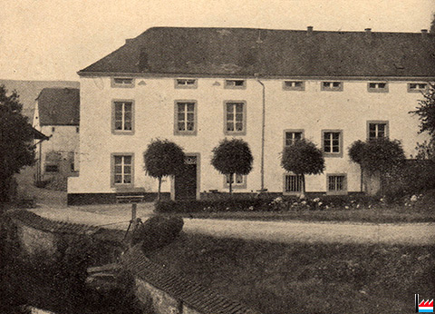 Une image sépia montre un bâtiment à Warken. Il semble idyllique avec quelques arbres au premier plan et son environnement villageois. Il s'agit de la première crique de Butzemillen, construite sur l'emplacement d'un ancien moulin (Millen en luxembourgeois) en 2013. Le moulin avait déjà été démoli en 1979.