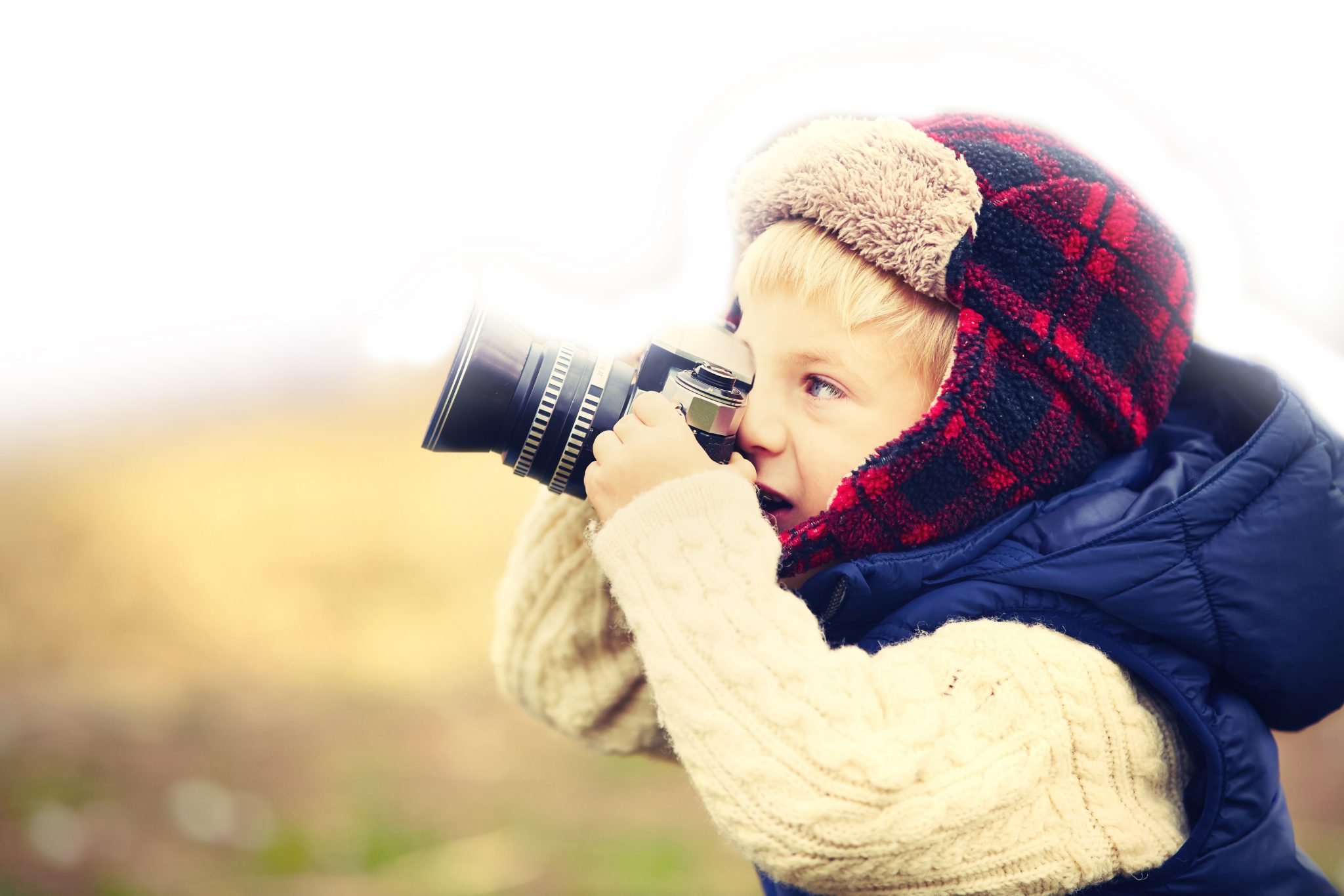 Un enfant blond avec un bonnet à carreaux rouges regarde dans un appareil photo en souriant.