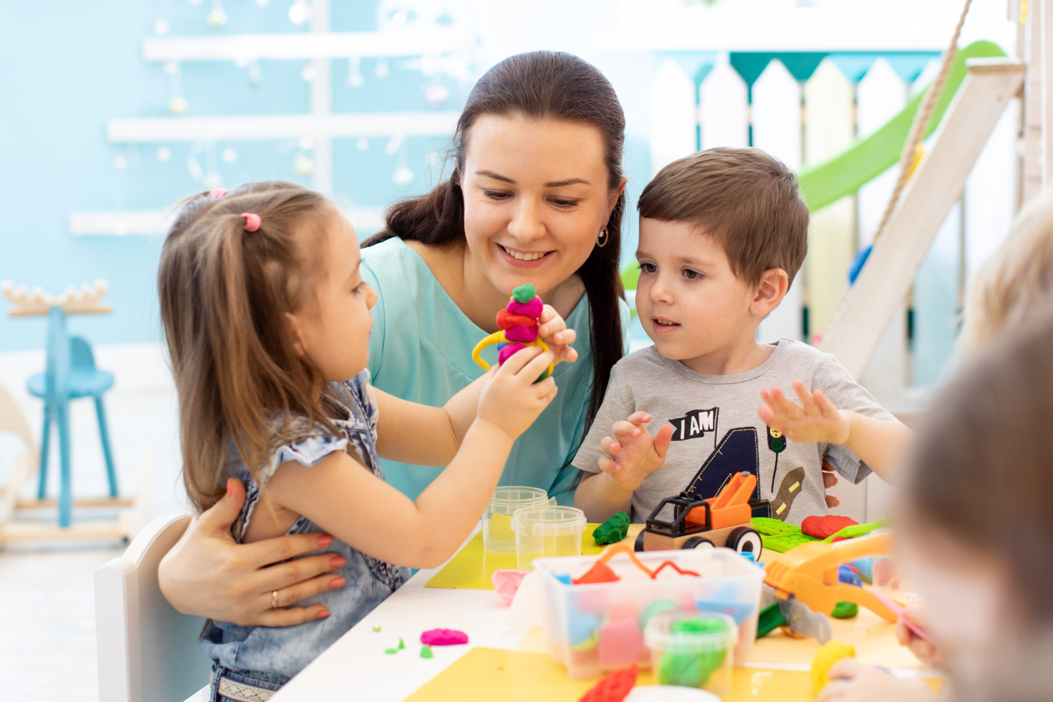 Une femme est assise à une table avec deux enfants, ils jouent avec des jouets colorés en pâte à modeler. En jouant ensemble avec nos éducateurs, il s'agit d'établir une profonde relation de confiance entre les enfants et nos animateurs. C'est essentiel pour une vie commune agréable dans nos crèches.